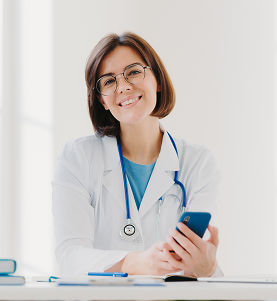A smiling person wearing a white coat and glasses holds a smartphone, with a stethoscope around their neck, sitting at a desk.